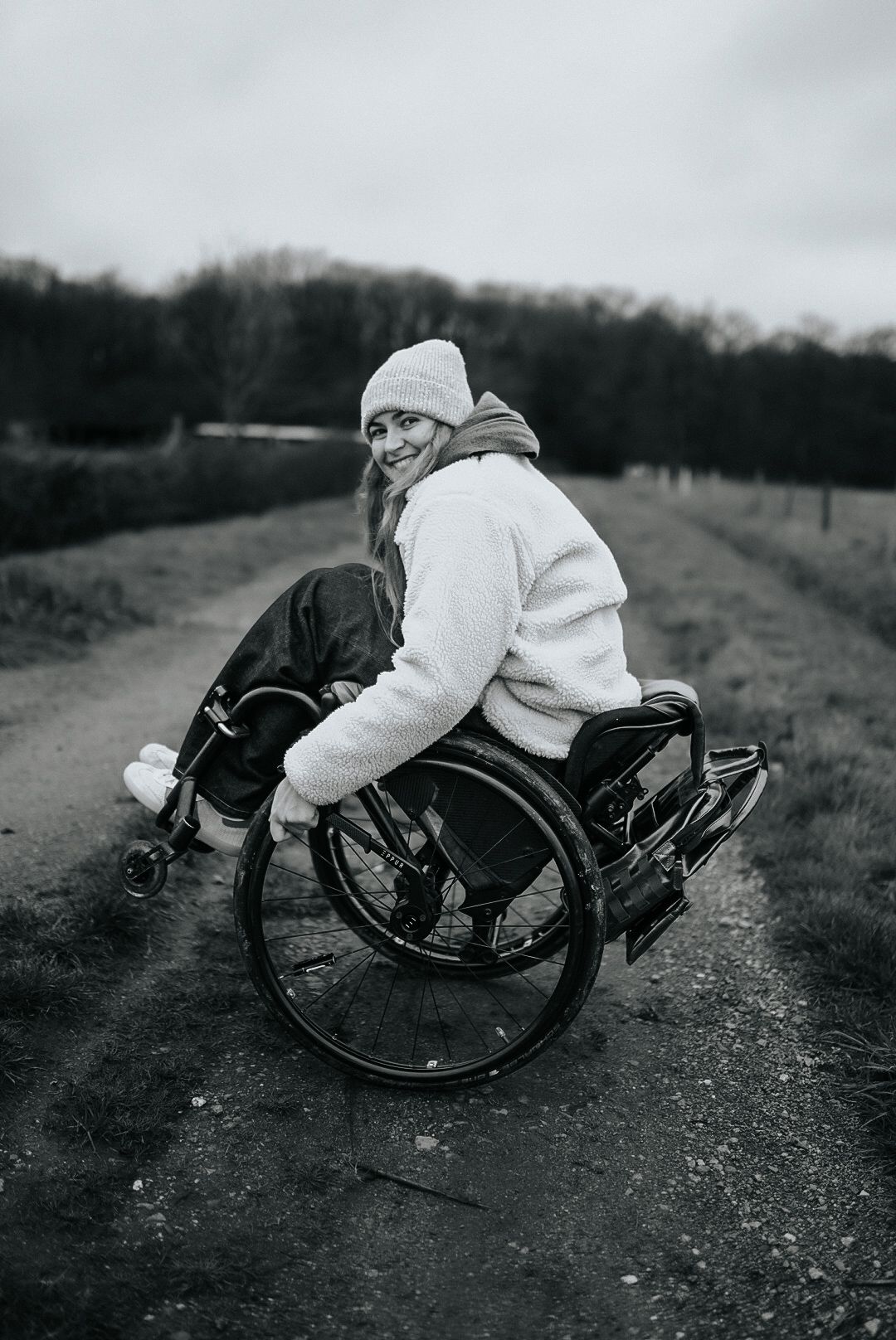 Femme souriante en fauteuil roulant sur un chemin rural portant une veste blanche moelleuse et un bonnet, photo en noir et blanc.