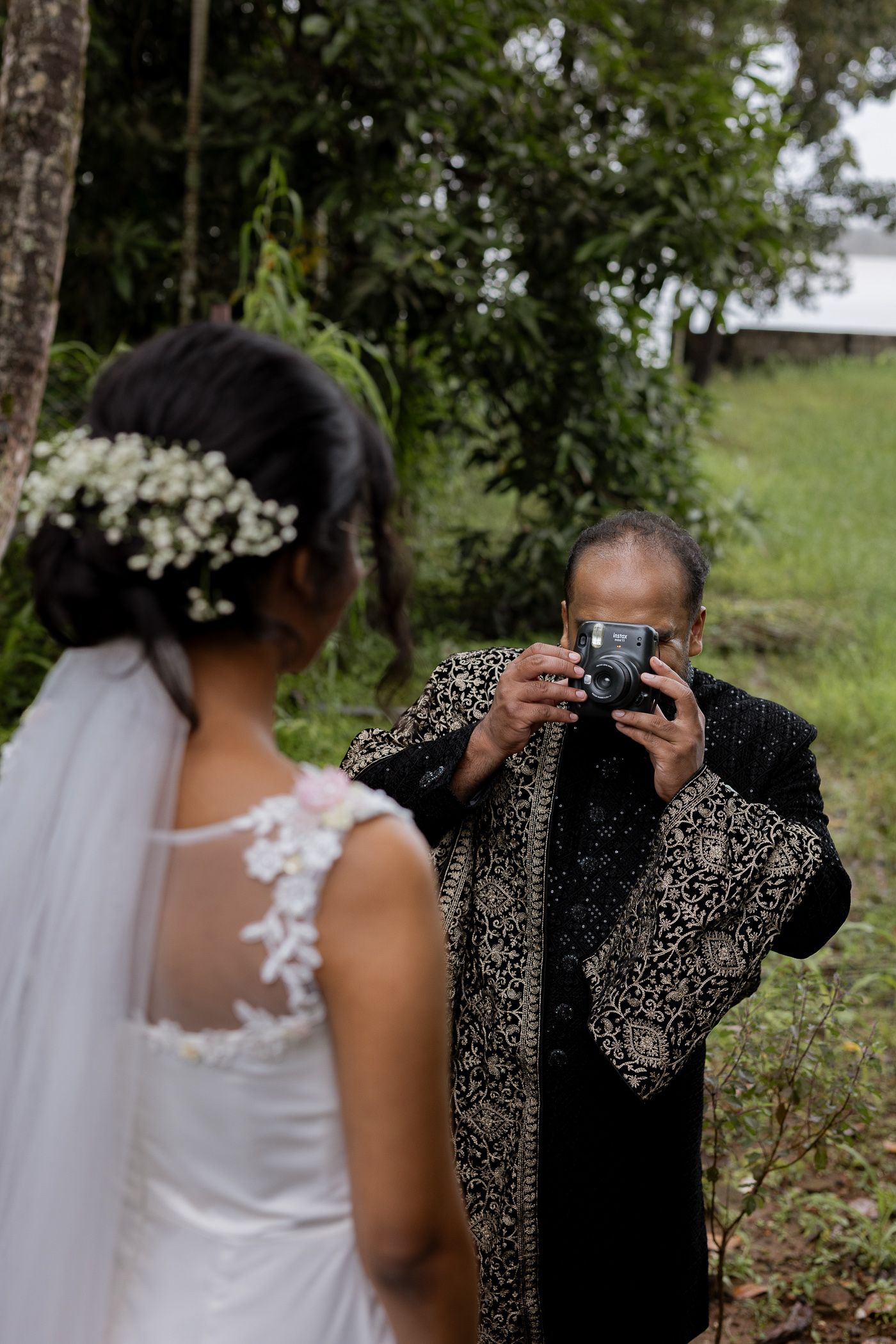 Bride in white lace gown with floral hair piece being photographed by man in ornate black suit outdoors.