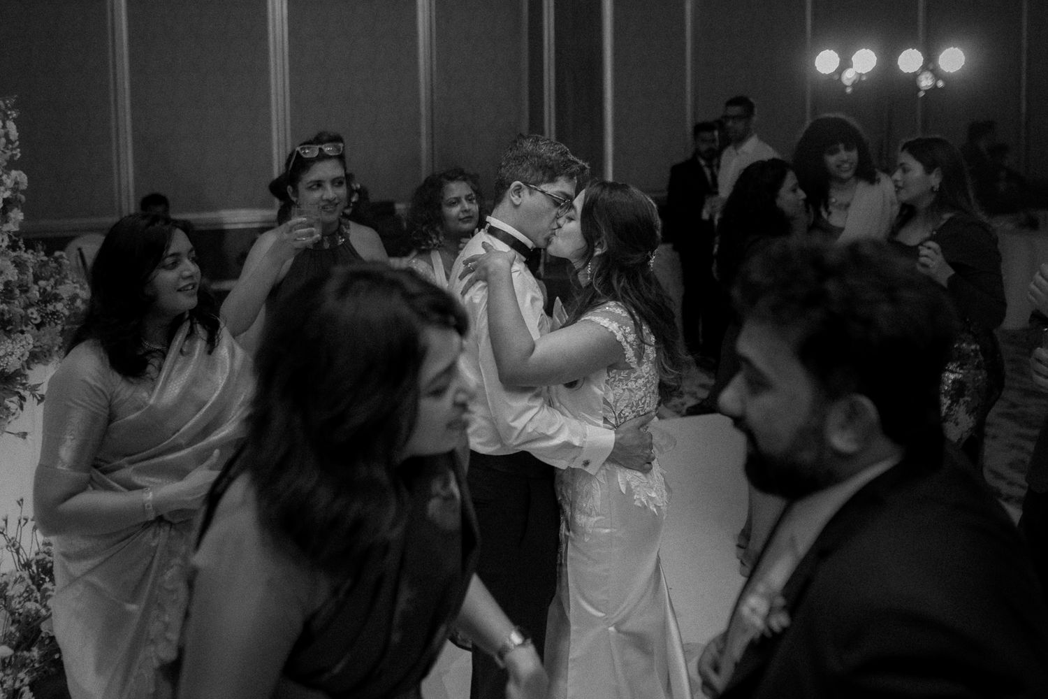Black and white wedding photo of couple sharing first dance surrounded by guests at elegant reception venue.