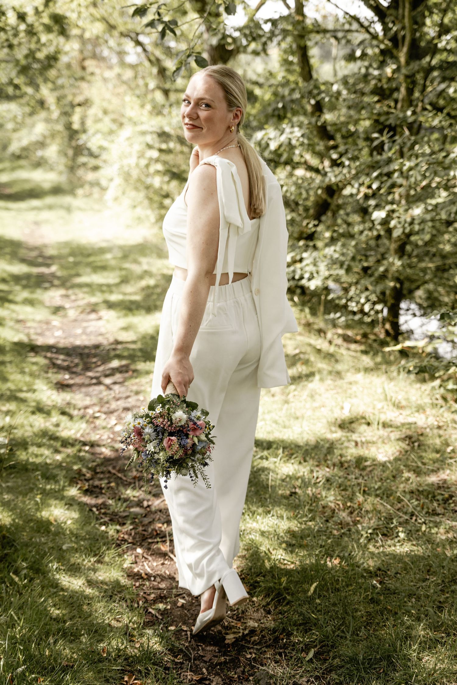 A bride in a white top and trousers holds a colorful bouquet while glancing back on a sunlit woodland path.