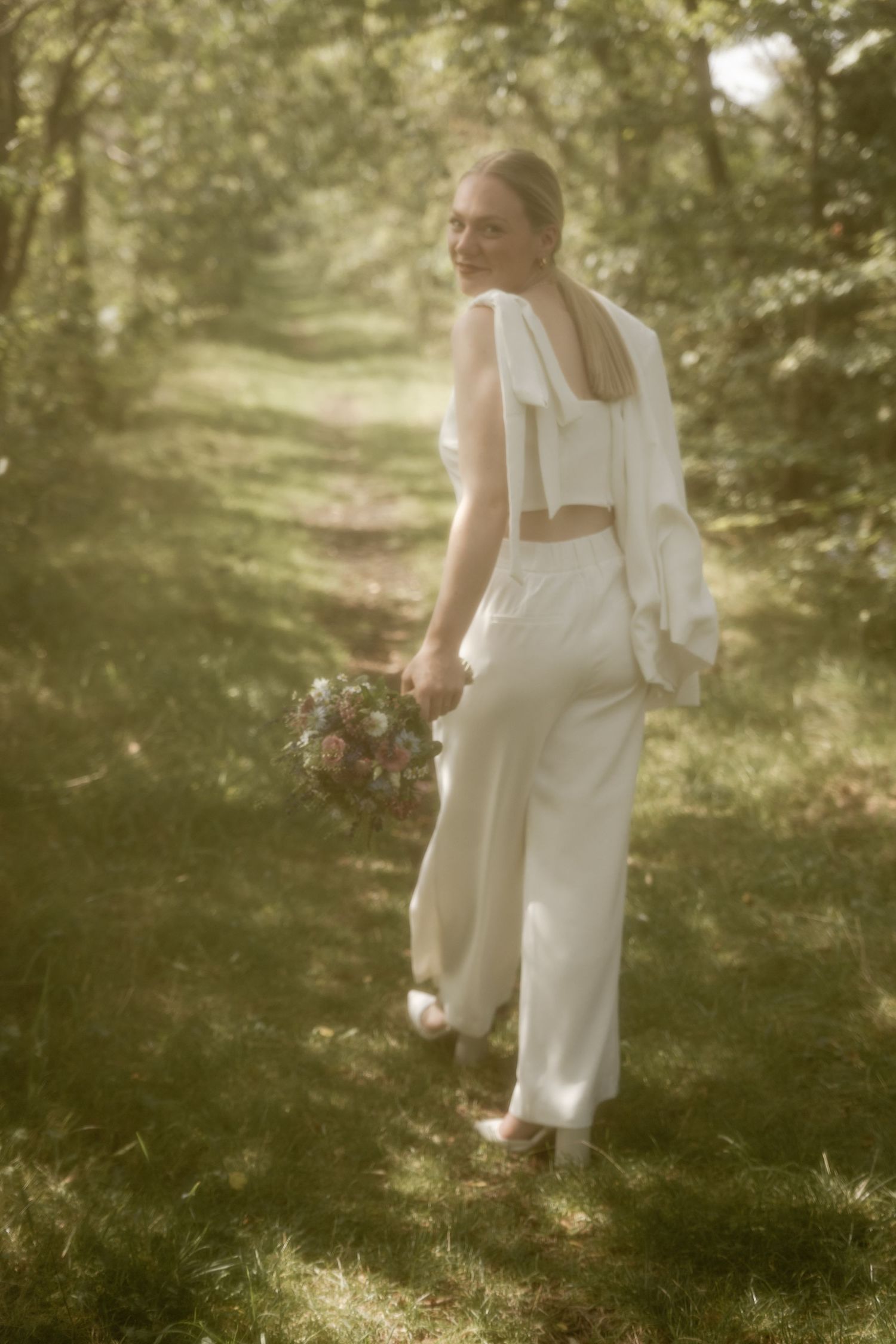 Bride in a white pantsuit with a bow-detailed back holds a bouquet while walking away along a sunlit woodland path.