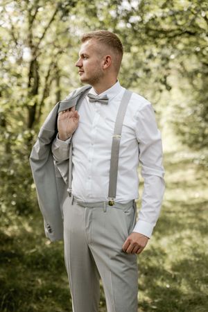 A groom in a white shirt, gray suspenders and bow tie holds his jacket over his shoulder while gazing aside in a green forest.