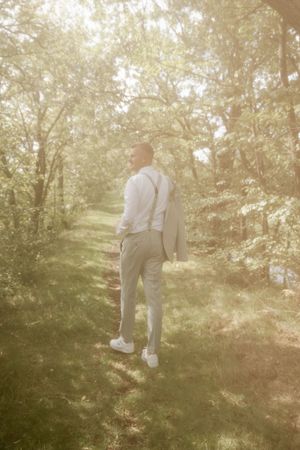Groom in gray trousers, white shirt and suspenders walks along a sunlit grassy path through the woods with his jacket over his shoulder.