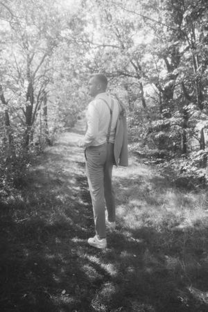 Black and white photo of a groom in suspenders carrying his jacket over his shoulder while standing on a wooded forest path.
