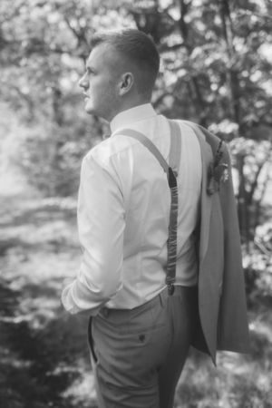 Black and white back view of a groom in a white shirt and suspenders with his jacket slung over his shoulder in the woods.