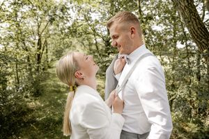 Smiling bride in a white blazer looks up lovingly at her groom as they embrace among green trees on their wedding day.