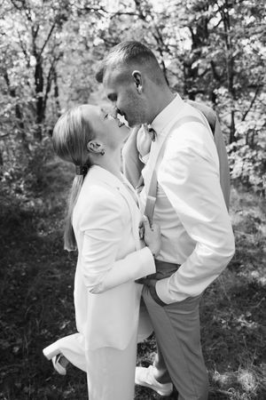 A black and white wedding photo of a bride and groom about to kiss in a leafy forest, her leg playfully lifted.