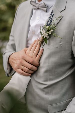 Close-up of a bride's hand with a wedding ring resting on a groom's chest, showing his gray suit, bow tie and floral boutonniere.