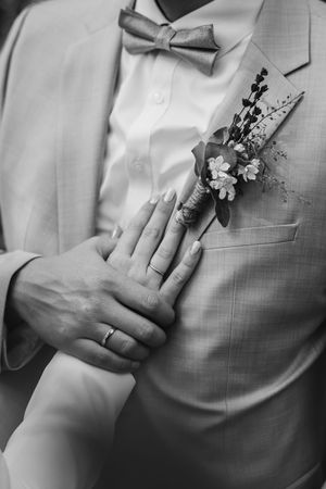 Black and white close-up of the bride's ringed hand on the groom's chest, with his bow tie and floral boutonniere in focus.