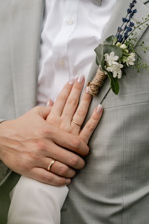 Close-up of a couple's hands with gold wedding rings resting on a groom's gray suit with a white floral boutonniere.