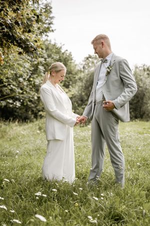 Bride in a white suit and groom in a light gray suit hold hands in a sunny meadow of daisies on their wedding day.