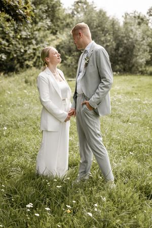 Bride in a white suit and groom in a light gray suit hold hands and gaze at each other in a sunny, flower-dotted meadow.