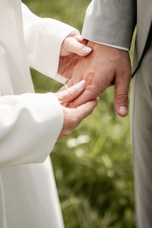 Close-up of the bride's hands holding the groom's hand showing her wedding band, with a soft green grassy background.