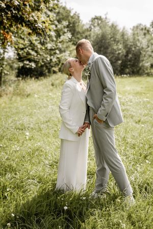 Groom in a gray suit kisses his bride in a white suit as they stand holding hands in a green summer meadow.