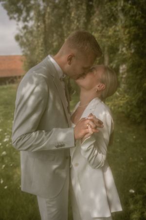 Bride and groom share a kiss outdoors on a grassy lawn, he in a light gray suit and she in a white blazer.