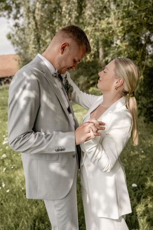 Smiling groom in a gray suit looks down at his bride in a white blazer as they hold hands among dappled sunlight.