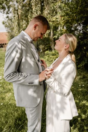 Groom and bride in white blazer share a tender look on green grass, holding hands during their outdoor wedding.