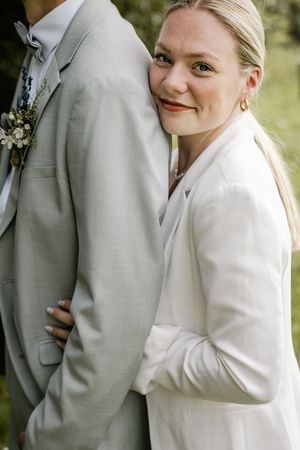 Smiling bride in a white blazer leans her head on the groom's shoulder, his gray suit adorned with a floral boutonniere.