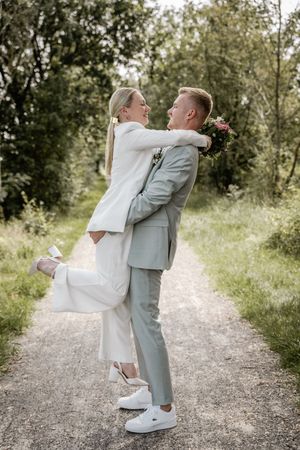 Groom in a gray suit and white sneakers lifts his laughing bride in a white suit on a gravel path surrounded by greenery.