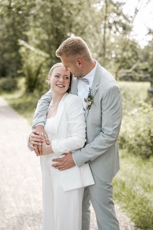 Happy bride in a white outfit smiles as the groom in a gray suit and bow tie embraces her from behind outdoors.