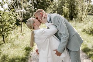Bride in a white blazer and ponytail kisses her groom in a gray suit on a sunny path lined with trees.