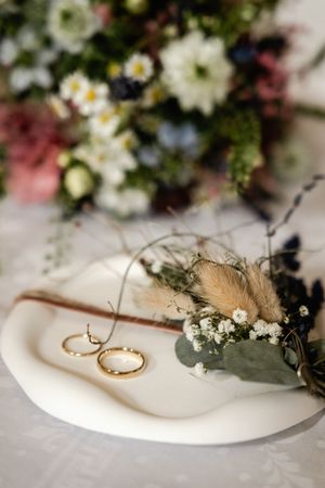 Two gold wedding rings rest on a white dish with dried pampas grass and small flowers beside a soft floral bouquet.