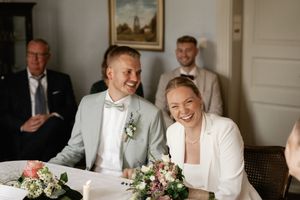 Smiling bride and groom sit at a table with a floral bouquet during their wedding ceremony as guests look on.