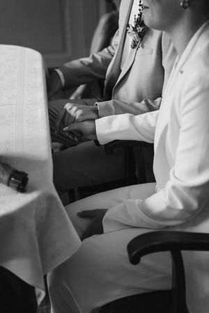 Black and white close-up of a bride and groom holding hands while seated at a lace-covered table during a wedding ceremony.