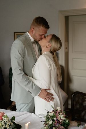 Groom in a sage green suit kisses his bride in a white blazer beside a bouquet of pink and white flowers.