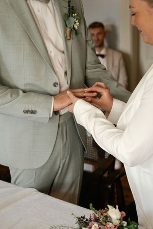 Braut im weißen Blazer steckt ihrem Bräutigam während der Trauung den Ehering an, während ein Hochzeitsgast zusieht. Museum Moorseer Mühle