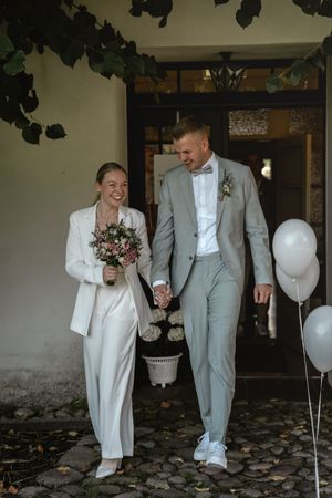 Frisch vermähltes Brautpaar hält Händche, verlässt die Trauung, während die Braut den Blumenstrauß trägt und Luftballons daneben schweben.