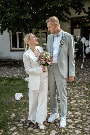Braut mit buntem Brautstrauß lächelt ihren Bräutigam im grauen Anzug in einem gepflasterten Innenhof nach der Hochzeit an.