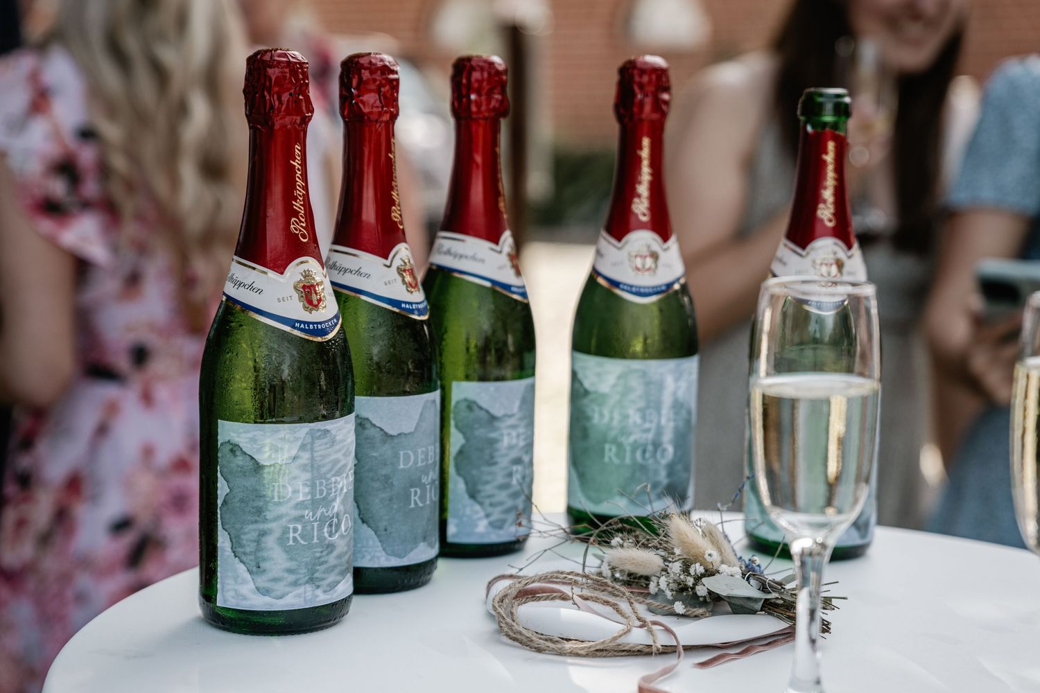 Flaschen mit personalisierten Etiketten und ein Champagnerglas stehen auf einem weißen Tisch bei einer Hochzeitsfeier.