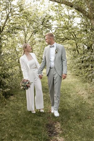 Bride in a white pantsuit holding a bouquet walks hand in hand with her groom in a gray suit along a grassy woodland path.