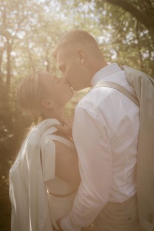 A bride and groom kiss in warm golden forest light, she in a white blazer and he in a shirt with suspenders.