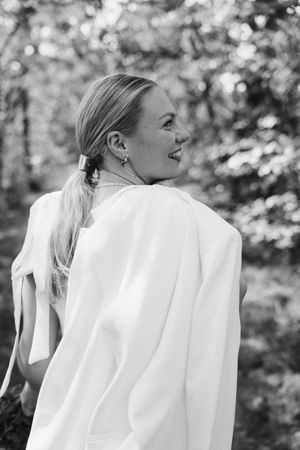 A black and white portrait of a smiling bride with a low ponytail and white blazer draped over her shoulders in a forest.