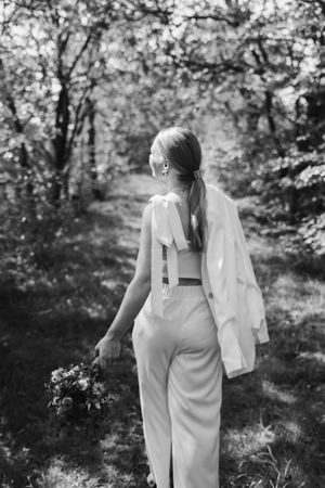 A black and white photo of a bride in a white jumpsuit holding a bouquet as she walks away along a wooded trail.