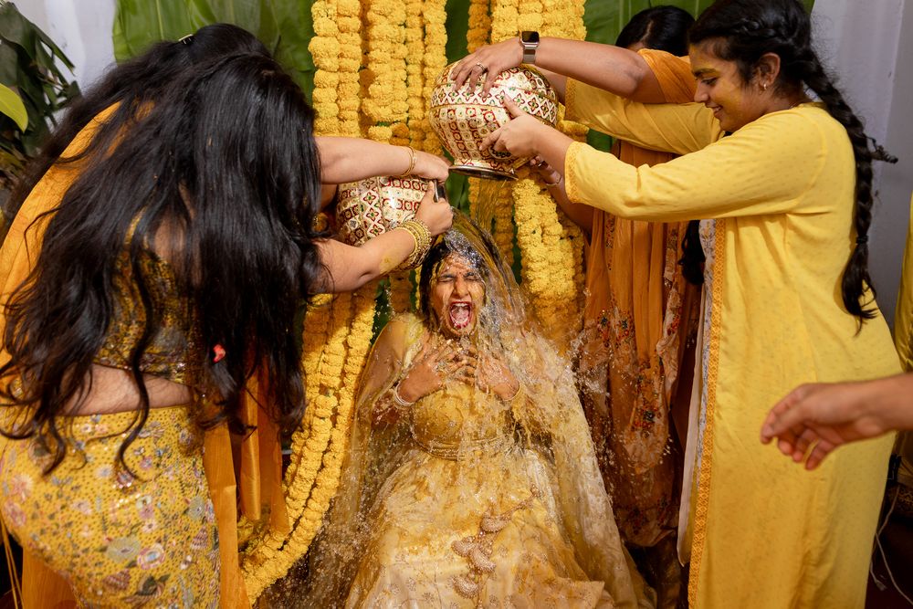 A bride in yellow laughs joyfully during a traditional Indian Haldi ceremony as friends pour turmeric paste over her.