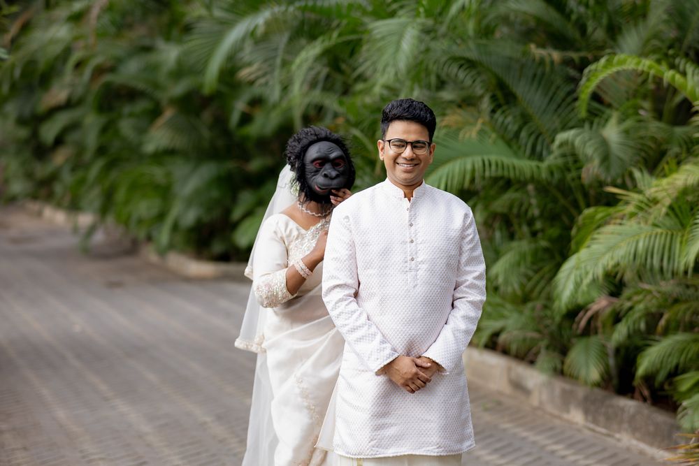 Indian couple in traditional white attire poses outdoors, groom in kurta smiles while bride holds decorative black mask.