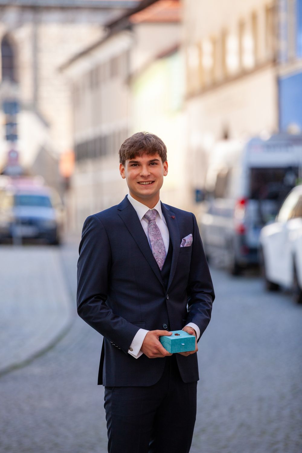 Young man in navy suit holding a teal gift box on a cobblestone European street.