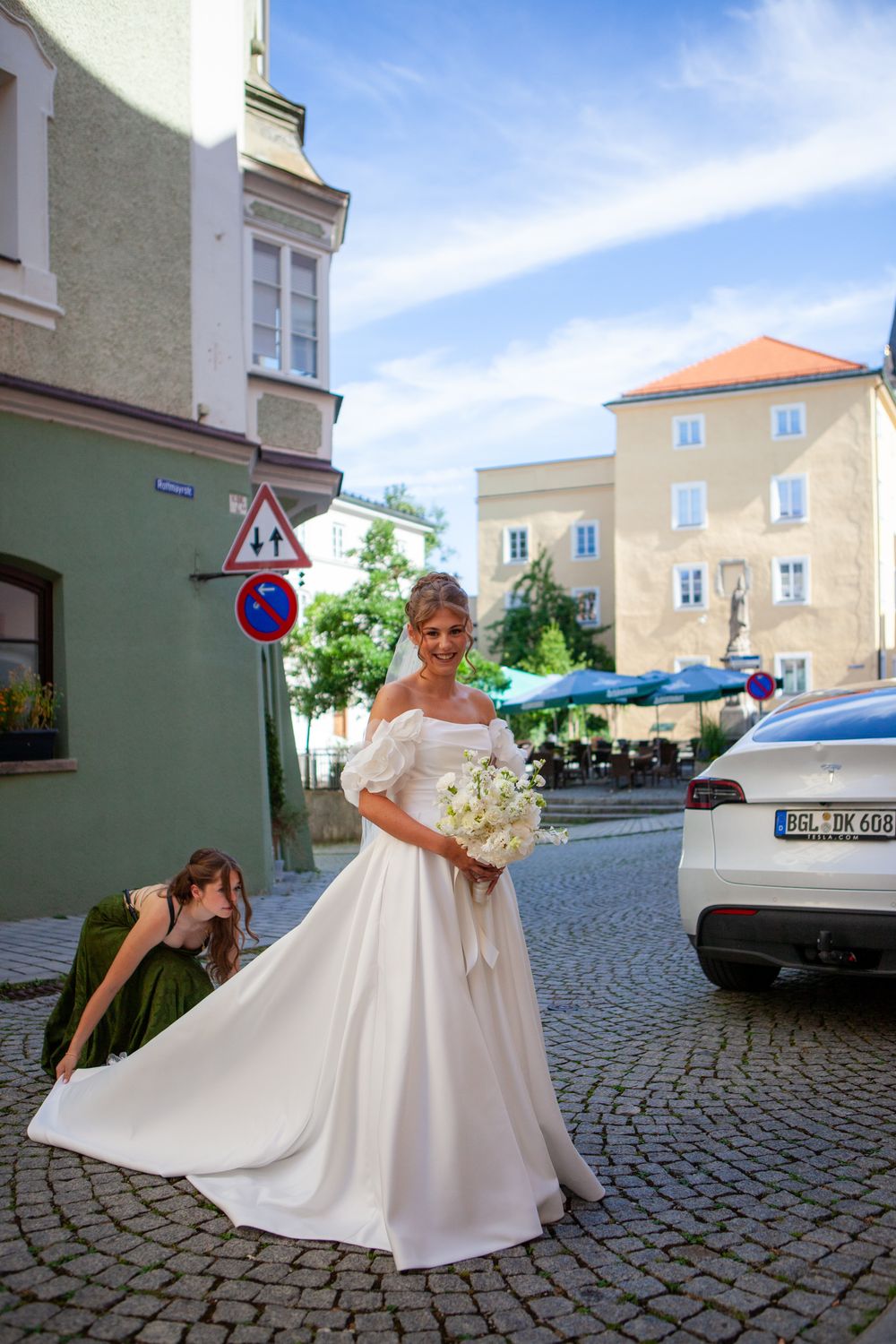 Bride in white off-shoulder gown holds bouquet on cobblestone street while bridesmaid adjusts her train.