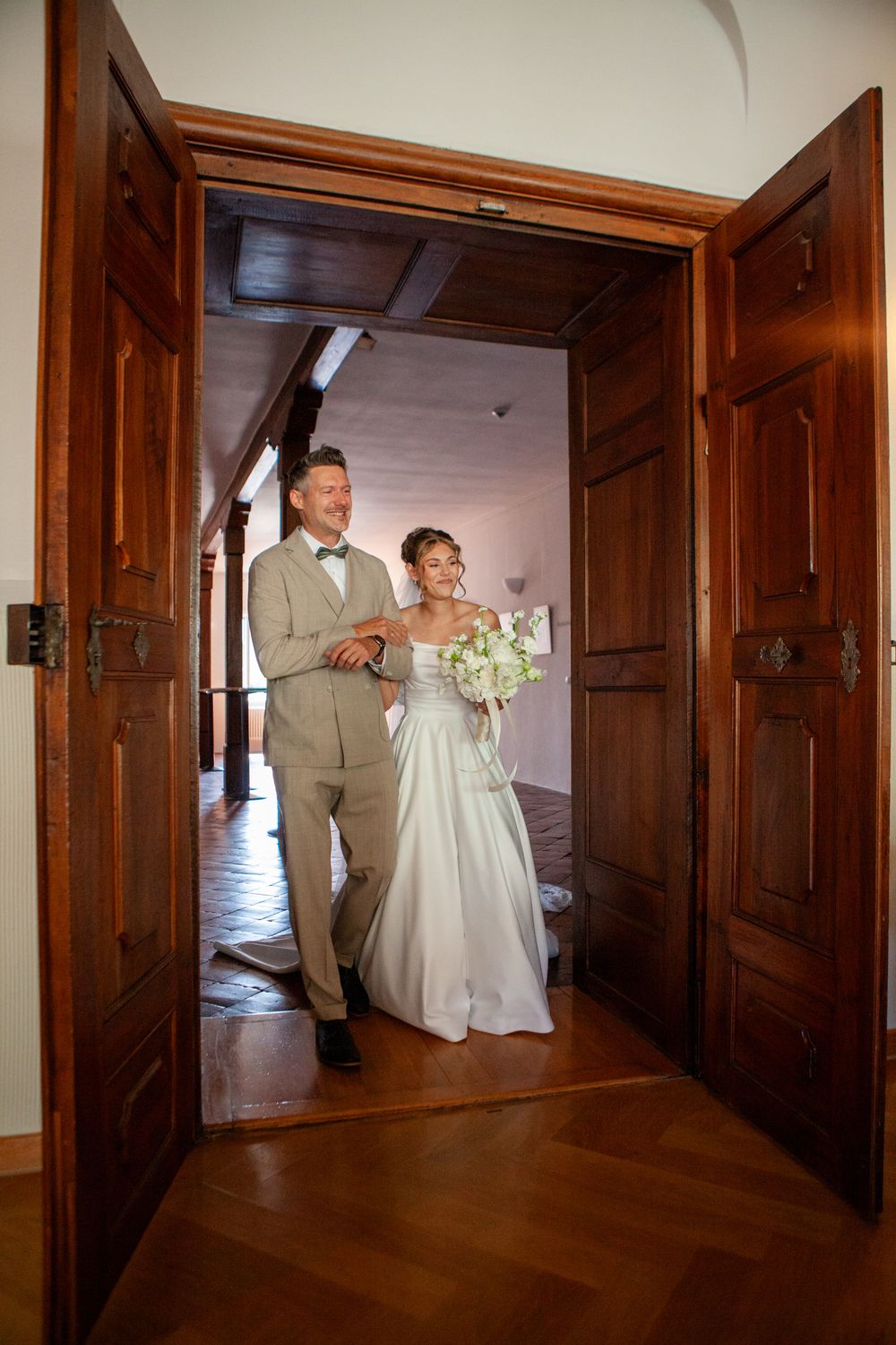 Smiling bride and groom walking through elegant wooden double doors at their wedding venue.
