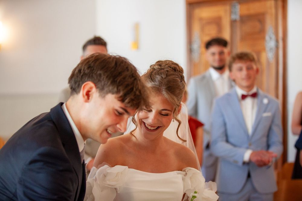 Smiling bride and groom share a joyful moment at their wedding ceremony with guests in formal attire behind them.