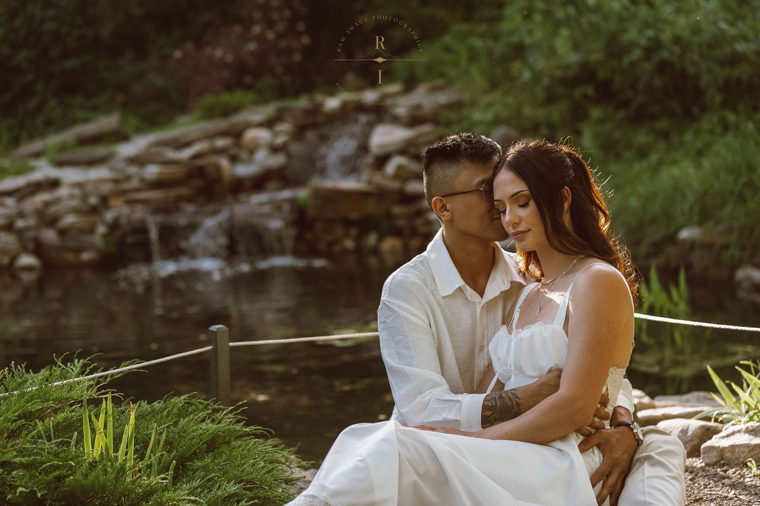 Couple in white attire sitting together by a serene garden waterfall surrounded by lush greenery.