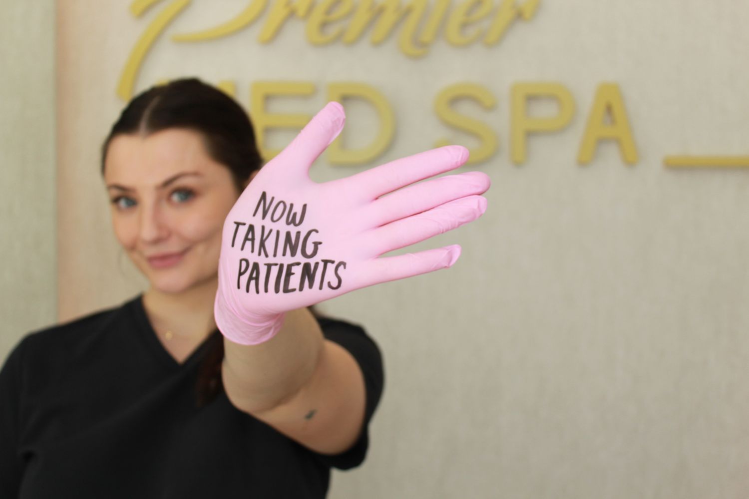 Woman holding pink glove reading Now Taking Patients at Premier Med Spa reception area.