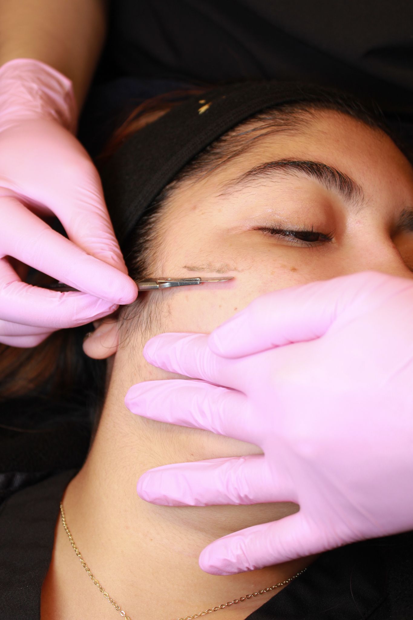 Esthetician in pink gloves performing precise eyebrow microblading treatment on a woman lying down.