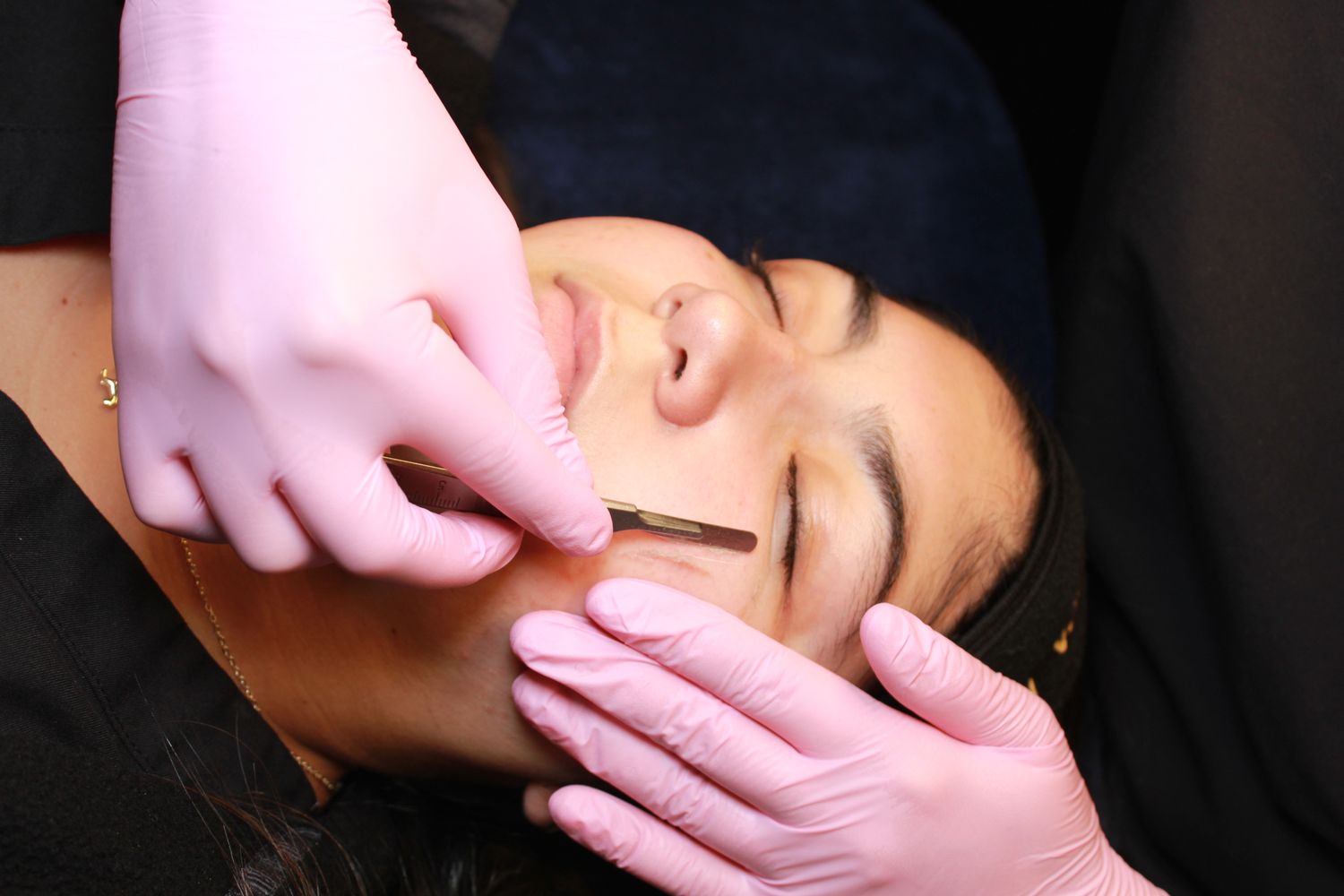 Esthetician in pink gloves performing a facial treatment on a woman lying down with eyes closed.