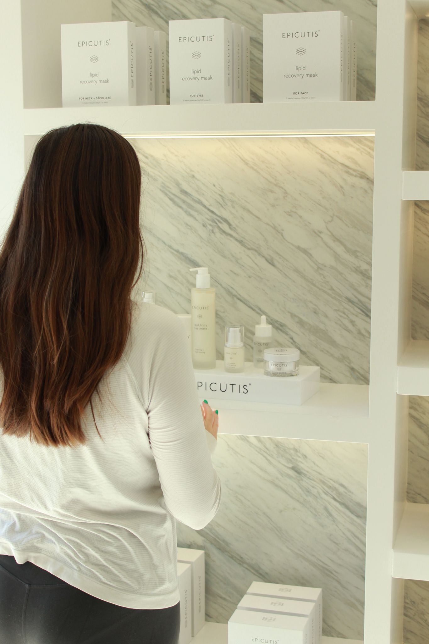Woman browsing Epicutis skincare products displayed on marble shelving unit in a luxury beauty retail store.
