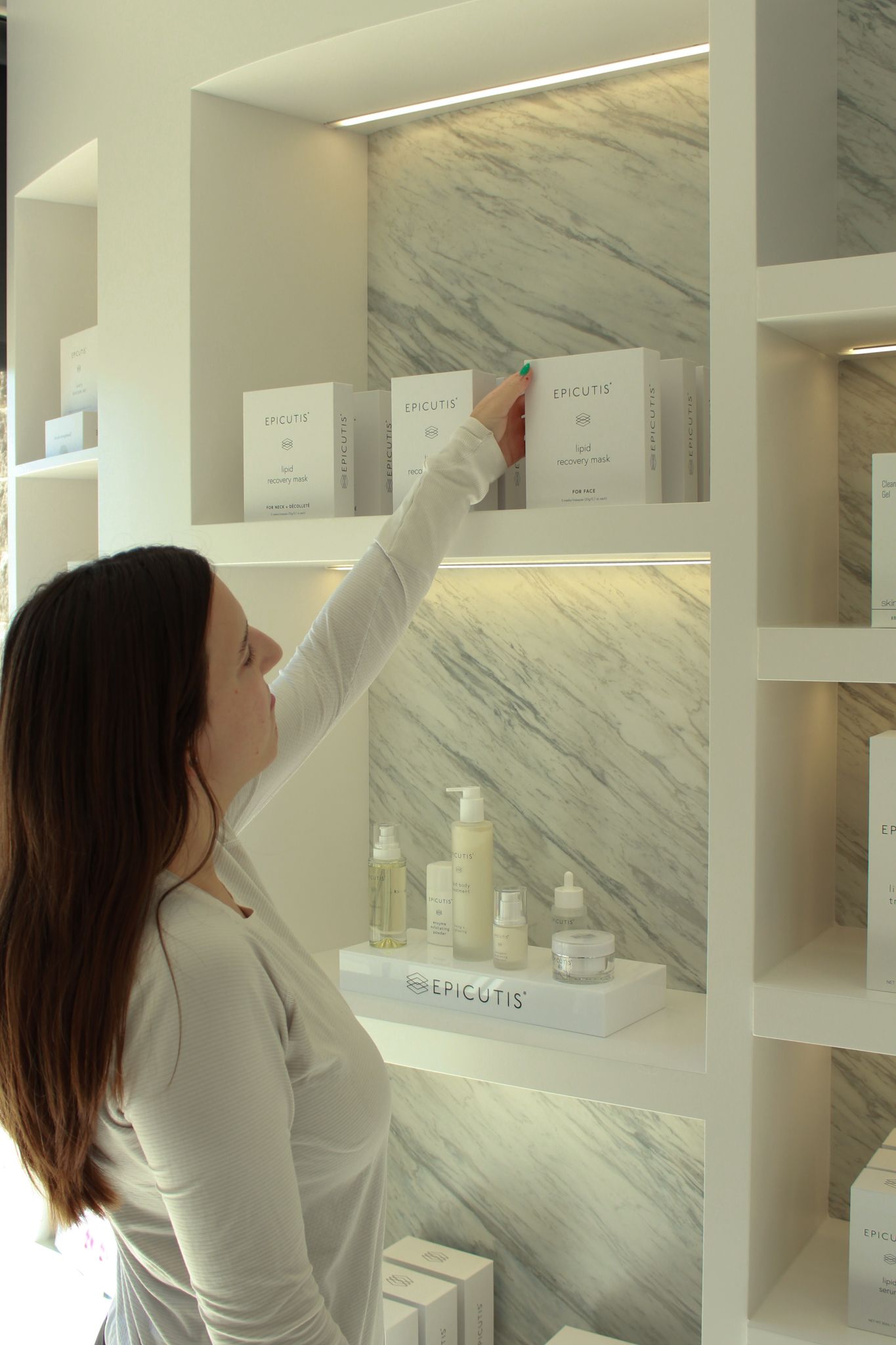 Woman reaching for Epicutis skincare boxes on illuminated marble shelves in an elegant beauty boutique.