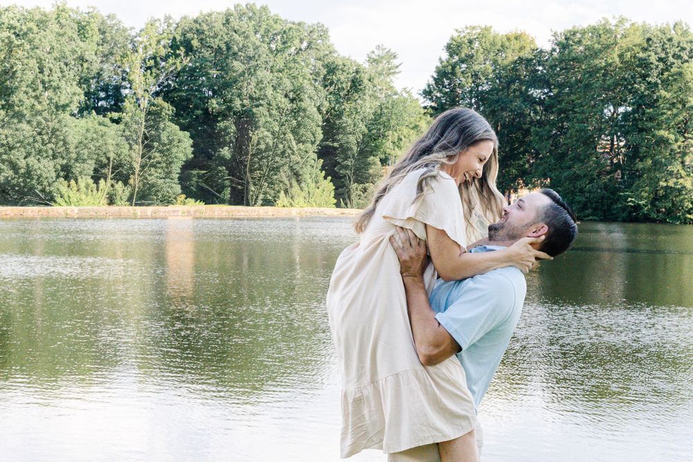 Couple sharing a romantic moment by a serene lake, man lifting woman in arms, surrounded by lush green trees.